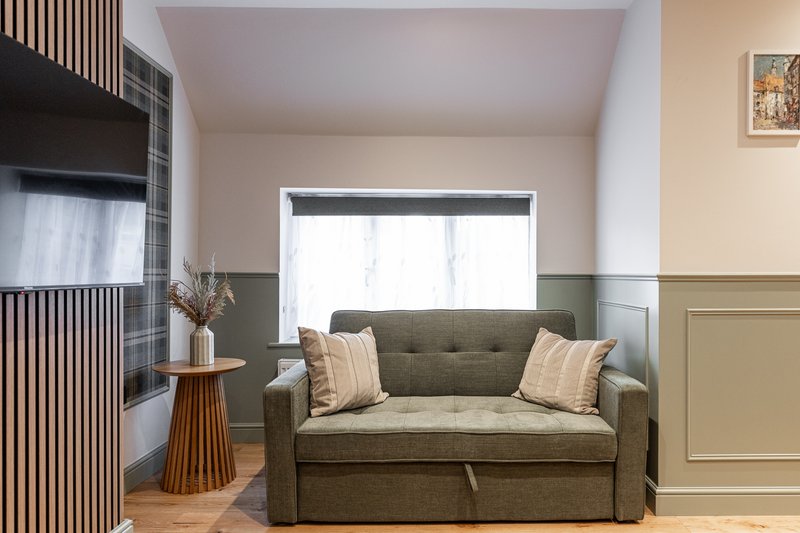 Modern living room with olive-green sofa, wooden side table, window with blind, and neutral wall panelling