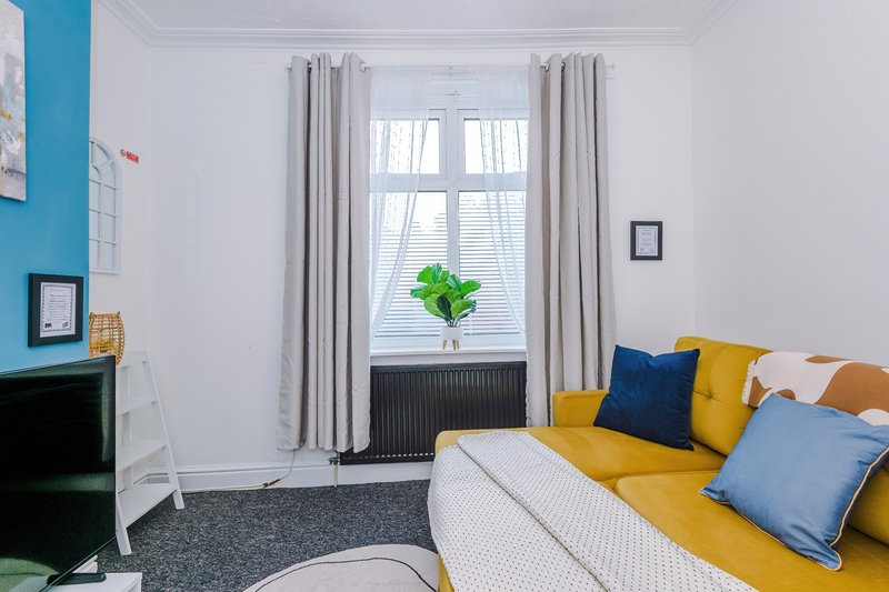 Bright bedroom with yellow sofa, blue and gold cushions, window with grey curtains and potted plant