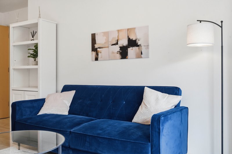 Modern living room with blue velvet sofa, white cushions, wall art, shelving unit and arc lamp