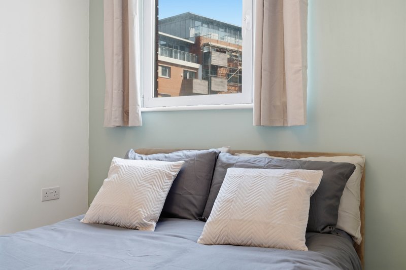 Bedroom with double bed, decorative pillows, window with curtains overlooking urban buildings