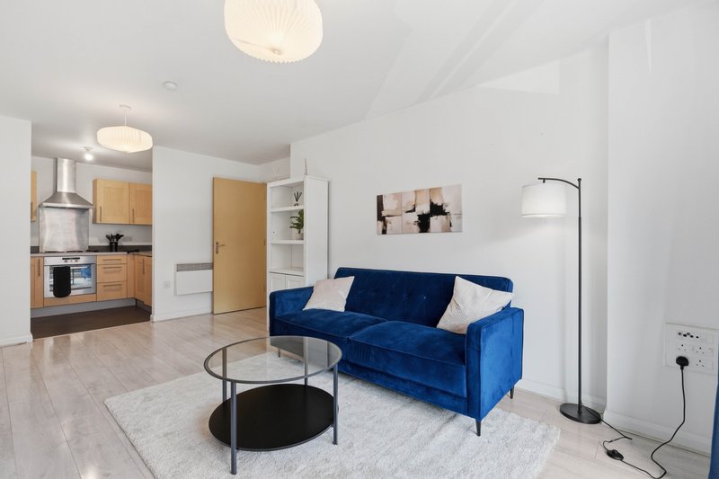 Modern open-plan living room with blue sofa, glass coffee table, kitchen area and floor lamp