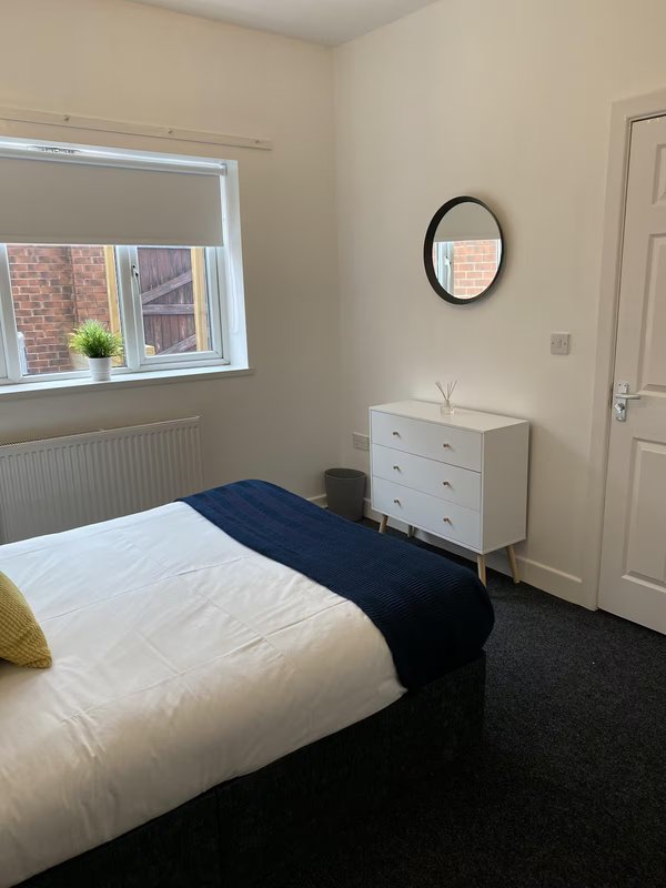 Bright bedroom with single bed, white dresser, oval mirror, and window overlooking brick building