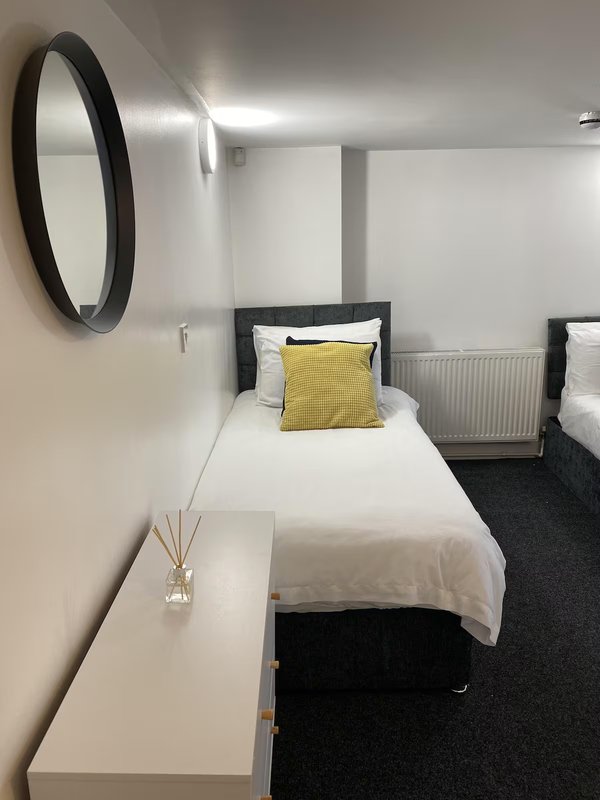 Bedroom with single bed, yellow cushion, round mirror, and white bedside table
