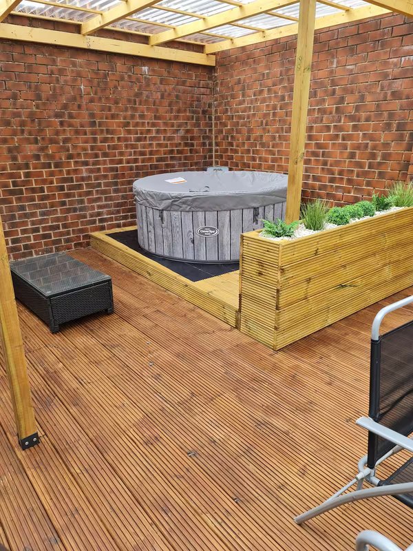 Covered outdoor patio with hot tub, wooden decking, brick walls, and seating area