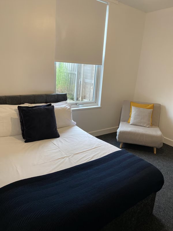 Bedroom with double bed, black headboard, cushions, and small upholstered chair by window