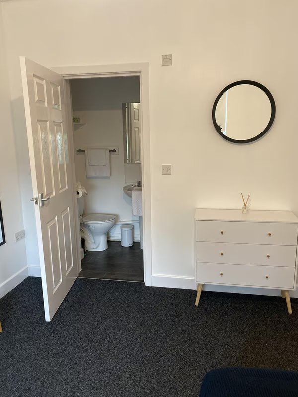 Bedroom corner with white dresser, round mirror, and open doorway to ensuite bathroom