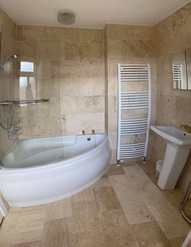 The en suite bathroom with curved white bathtub, glass shower enclosure, heated towel rail and pedestal sink