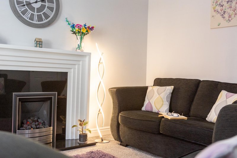 Living room with brown sofa, fireplace, wall clock, colorful flowers in vase, and ambient lighting