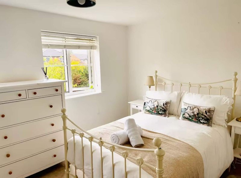 Bright bedroom with brass bed frame, white bedding, cream dresser and window with garden view