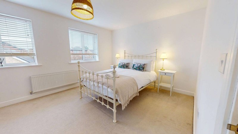 Bright bedroom with double bed, metal frame, two windows, bedside lamps and carpet flooring