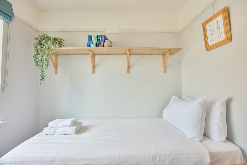 Bright bedroom with double bed, white bedding, wooden wall shelf with plants and books