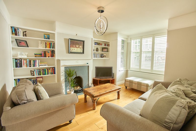Bright living room with two cream sofas, fireplace, bookshelves, wooden coffee table and shuttered windows