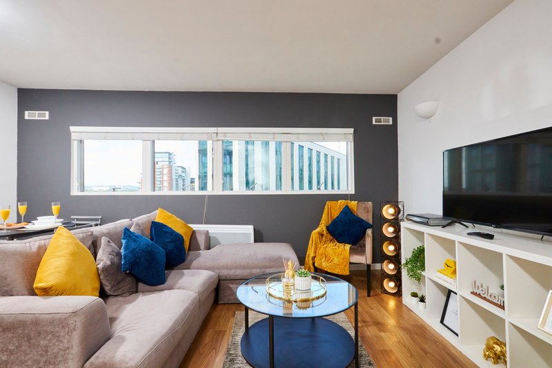 Modern living room with beige sofa, yellow and blue cushions, TV, and city view through window