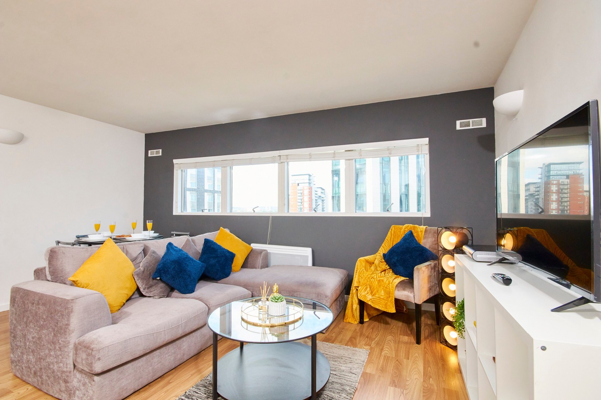 Modern living room with gray sofa, yellow and blue cushions, glass coffee table and city views