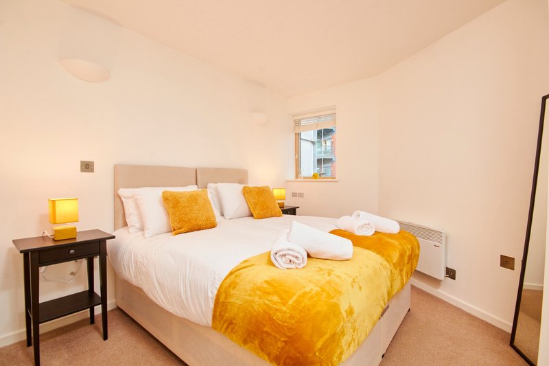 Bright bedroom with double bed, yellow velvet bedding, white pillows, wooden bedside table and window