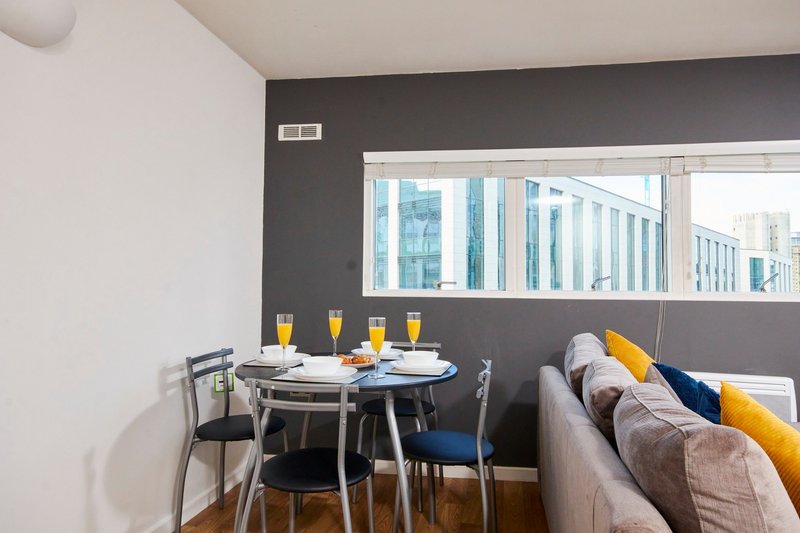 Modern open-plan living area with dining table, chairs, sofa with cushions, and city views through windows