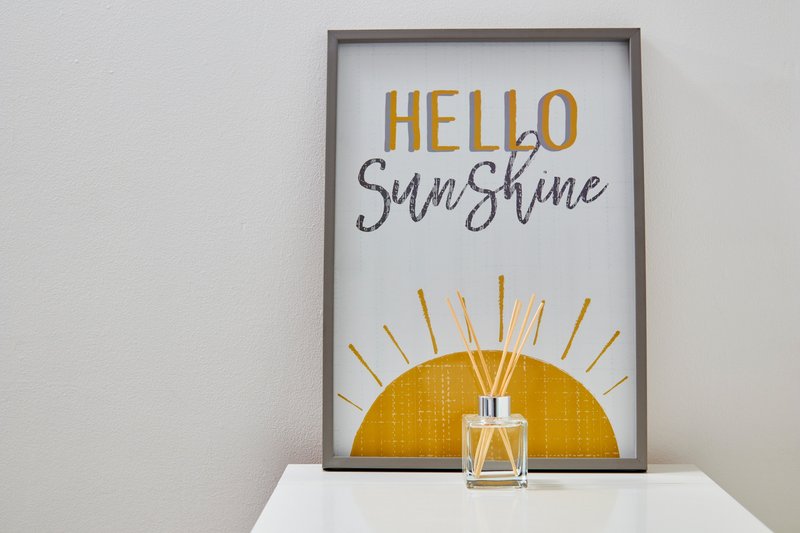 Framed decorative print reading 'Hello Sunshine' with yellow sun illustration and perfume bottle on white shelf