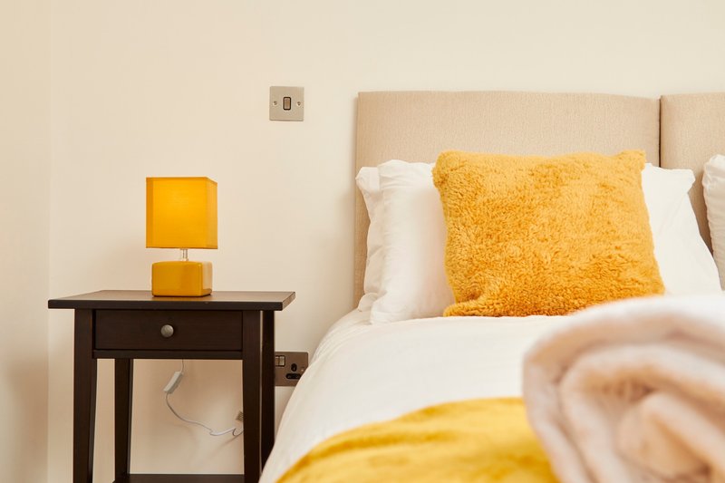 Bedroom with beige upholstered headboard, yellow cushion, white pillows, dark wooden bedside table with yellow lamp