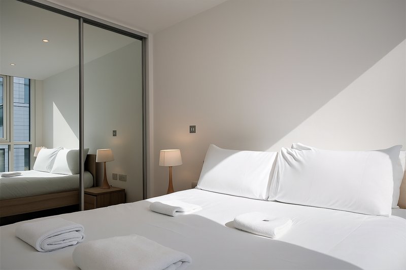 Modern bedroom with double bed, mirrored wardrobe, bedside lamp and white linens