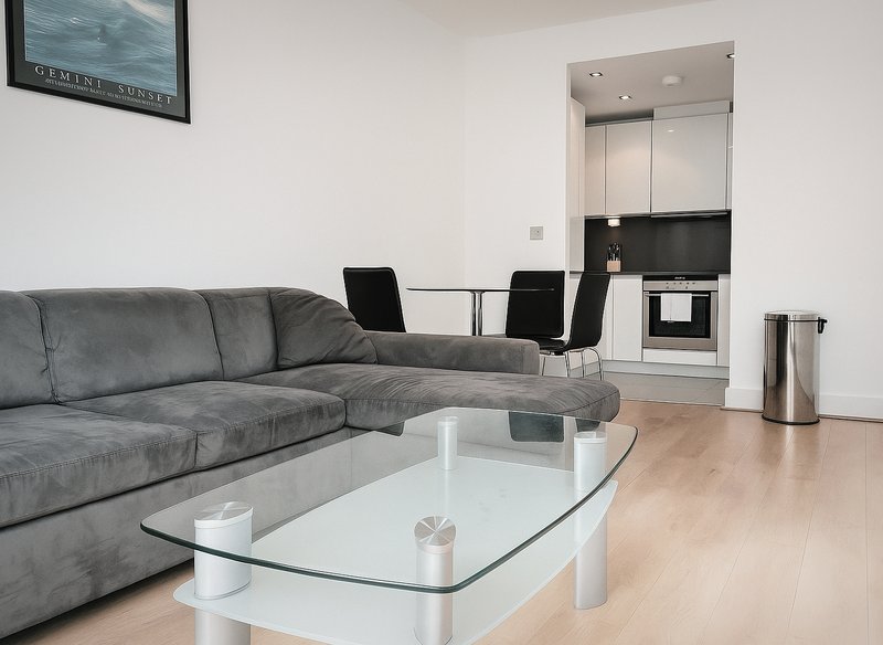 Modern living room with grey corner sofa, glass coffee table, and open-plan kitchen area