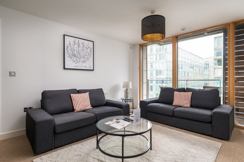Modern living room with two black sofas, round glass coffee table, framed wall art, and large windows overlooking city buildings.