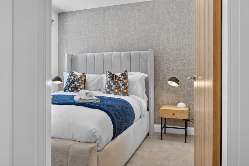 Modern bedroom with upholstered headboard, white bedding, blue throw, patterned cushions and wooden nightstand