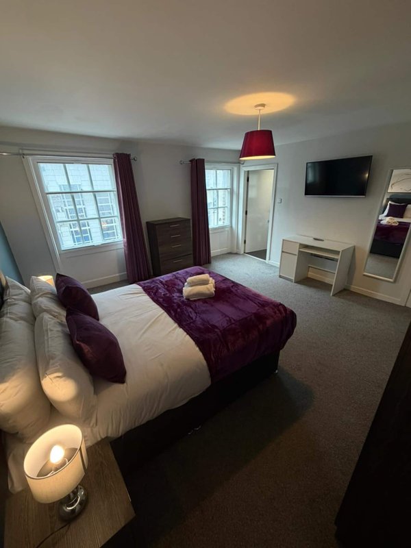 Spacious bedroom with double bed, purple bedding, TV, and dual windows with curtains
