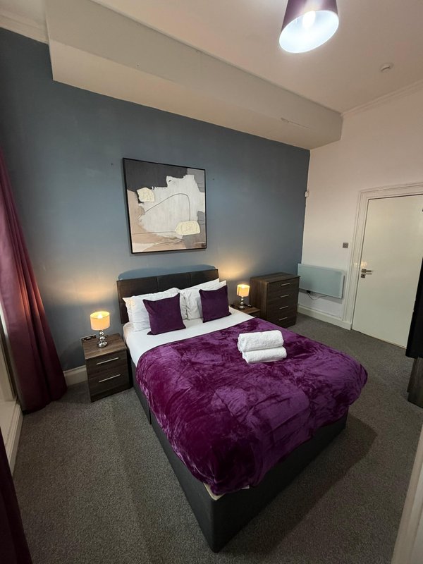 Modern bedroom with purple duvet, dark headboard, bedside lamps, and blue accent wall