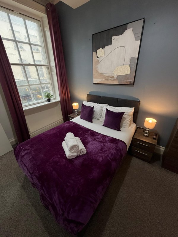 Bedroom with purple velvet bed, dark headboard, window with burgundy curtains, wall art, and bedside lamps