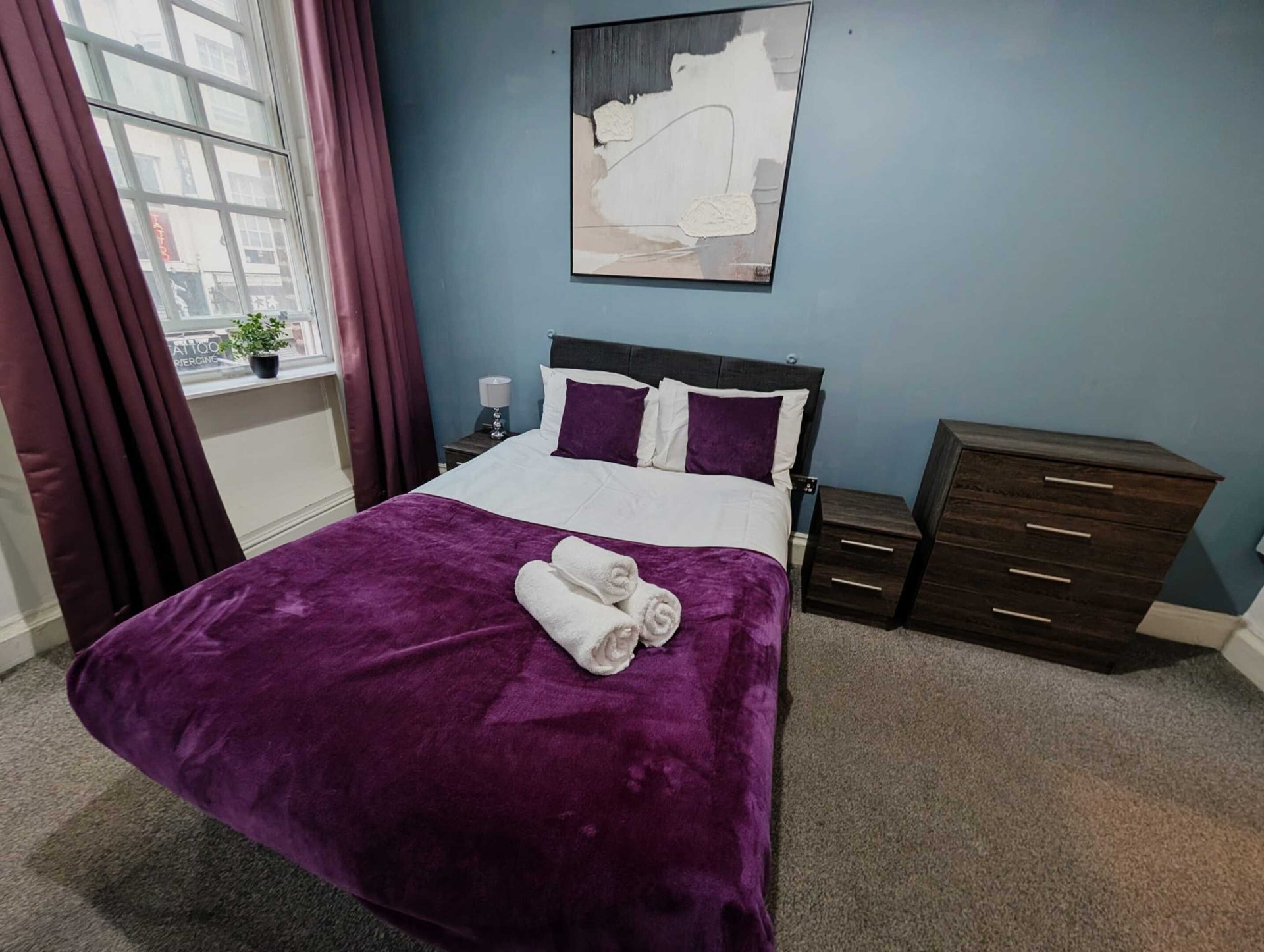 Bedroom with purple velvet bedspread, white rolled towels, dark wood dresser and blue walls