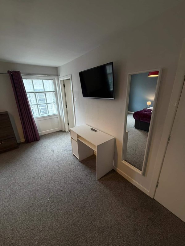 Bright apartment living room with wall-mounted TV, white desk, mirror, and window with burgundy curtains