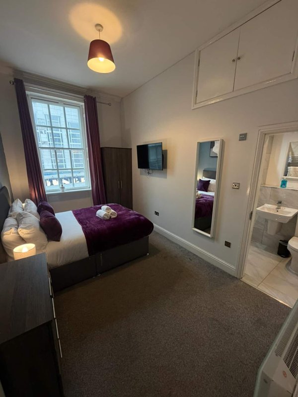 Modern bedroom with double bed, purple bedding, window with curtains, and ensuite bathroom visible