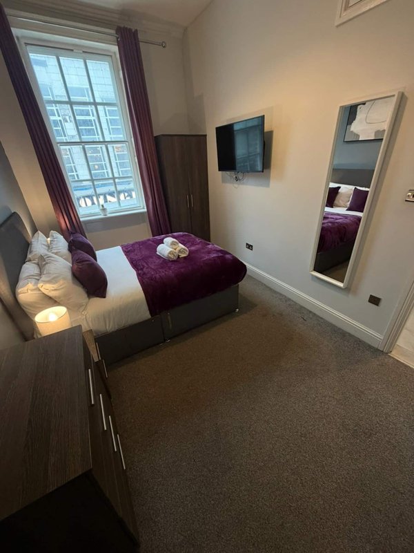 Compact bedroom with double bed, purple bedding, wall-mounted TV, mirror and window with curtains