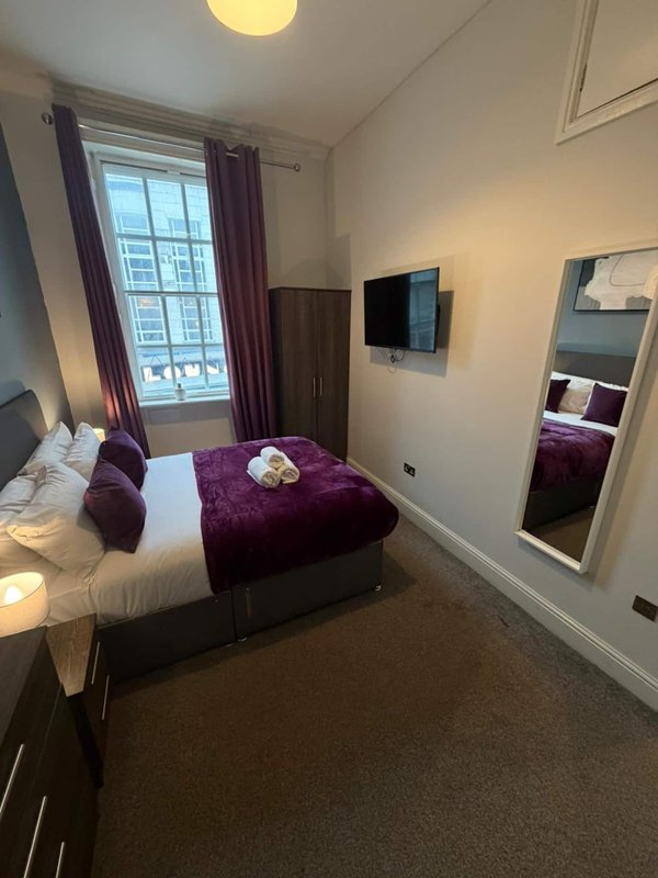Compact bedroom with single bed, purple bedding, wall-mounted TV and large window with curtains