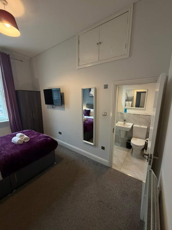 Bedroom with purple bed, wall-mounted TV, en-suite bathroom door, and burgundy curtains