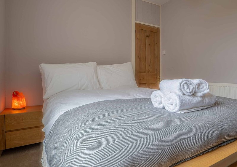 Bedroom with double bed, white pillows, gray bedding, rolled towels, wooden door and bedside lamp
