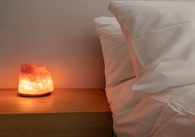 Bedroom with rumpled white bedding and glowing salt lamp on wooden nightstand