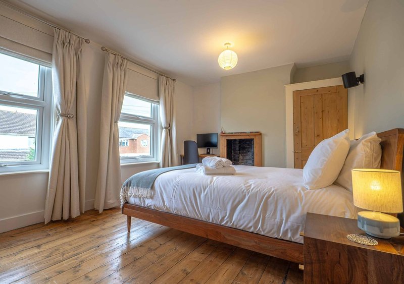 Bright bedroom with wooden bed, white bedding, large windows with cream curtains, wooden floor