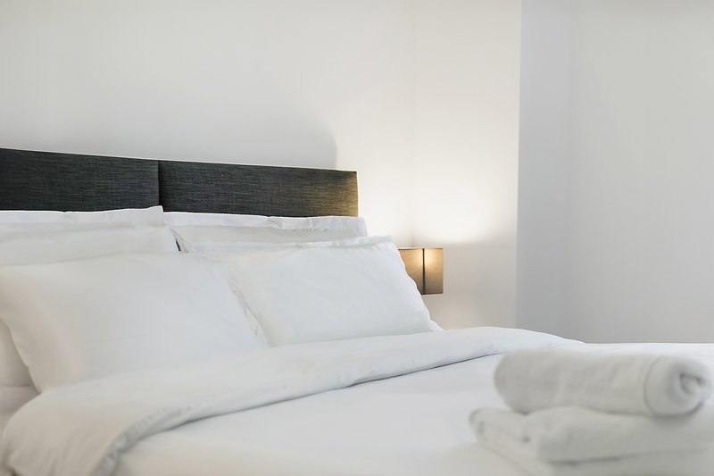 Modern bedroom with white bedding, dark headboard, bedside lamp and rolled towels