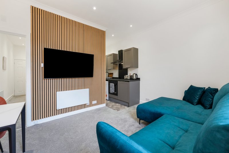 Modern living room with turquoise sofa, wall-mounted TV, wooden feature wall, and kitchenette