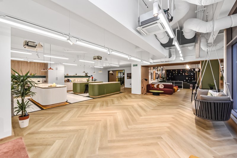 Modern open-plan office or communal workspace with herringbone flooring, kitchen area, seating zones, and exposed ceiling systems