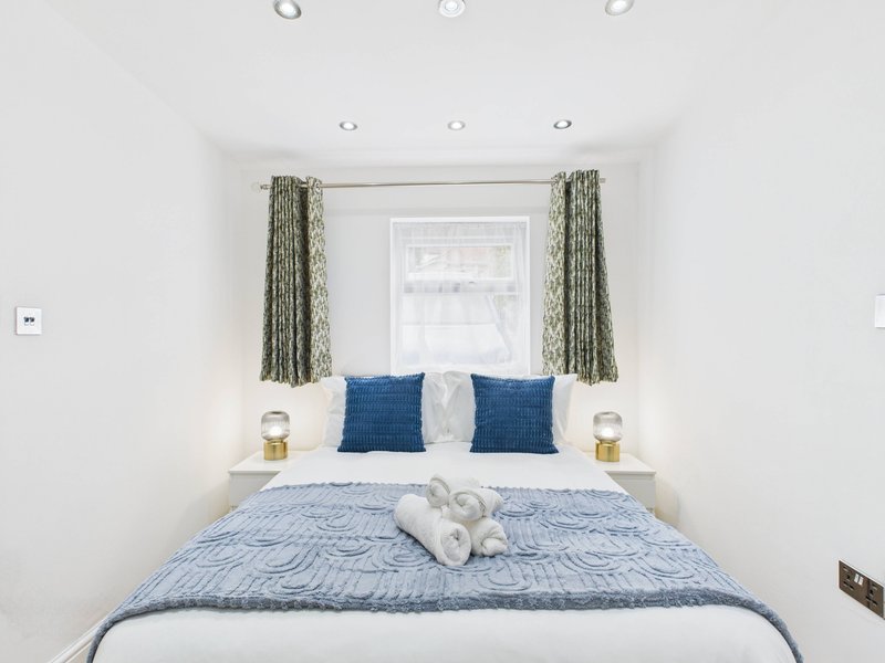 Bright bedroom with double bed, blue bedding, navy cushions, green curtains and bedside lamps
