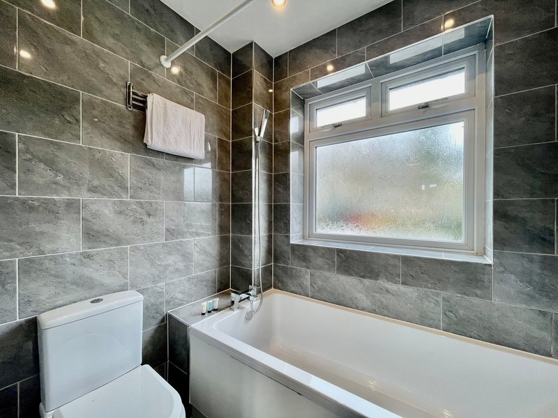 Modern bathroom with grey tile walls, white bathtub, toilet, and window with natural light