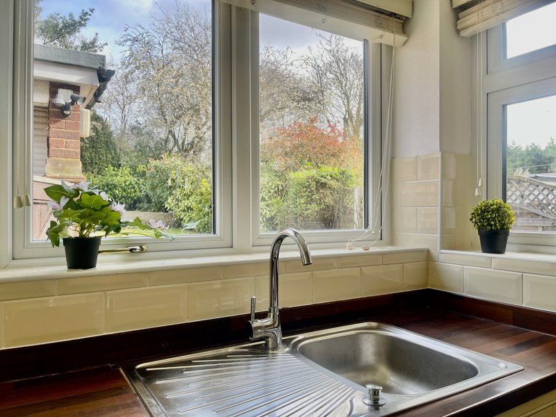 Kitchen sink with stainless steel basin, tap and drainboard, cream tile splashback, large windows with garden views and potted plants on sill