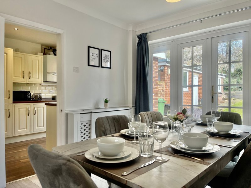 Dining room with laid table, kitchen visible through doorway, large windows with garden view