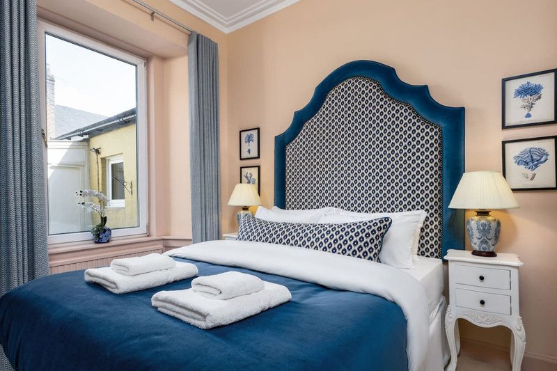 Bedroom with blue upholstered headboard, white bedding, blue throw, nightstand lamp and window