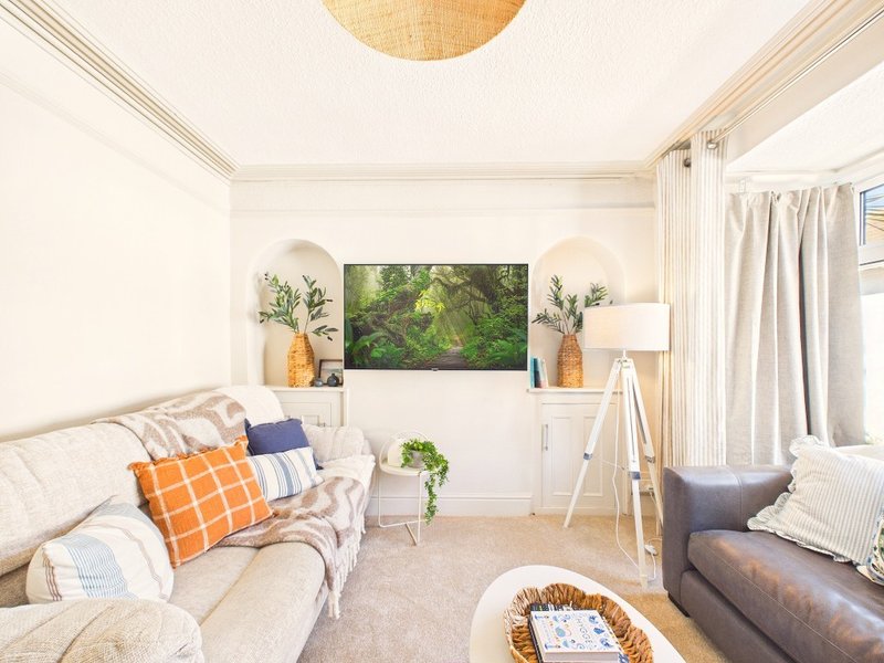 Bright living room with cream sofa, wall-mounted TV, indoor plants, and natural lighting from windows
