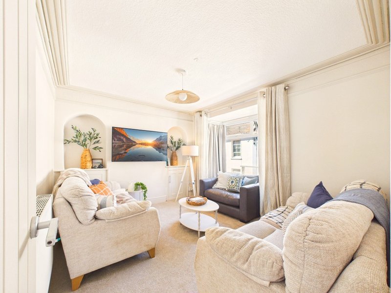 Bright living room with two beige sofas, framed landscape artwork, potted plants, and natural light from windows