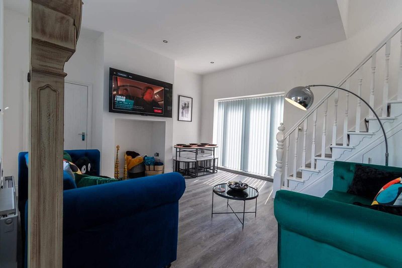 Open-plan living room with blue and green sofas, mounted TV, stairs, and natural light from patio doors
