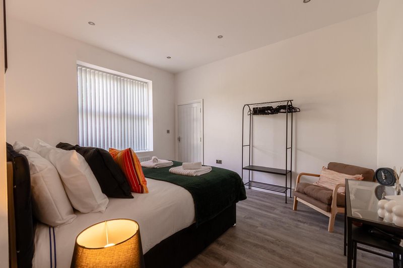 Spacious bedroom with double bed, cushions, bedside lamp, clothing rack, and window with blinds